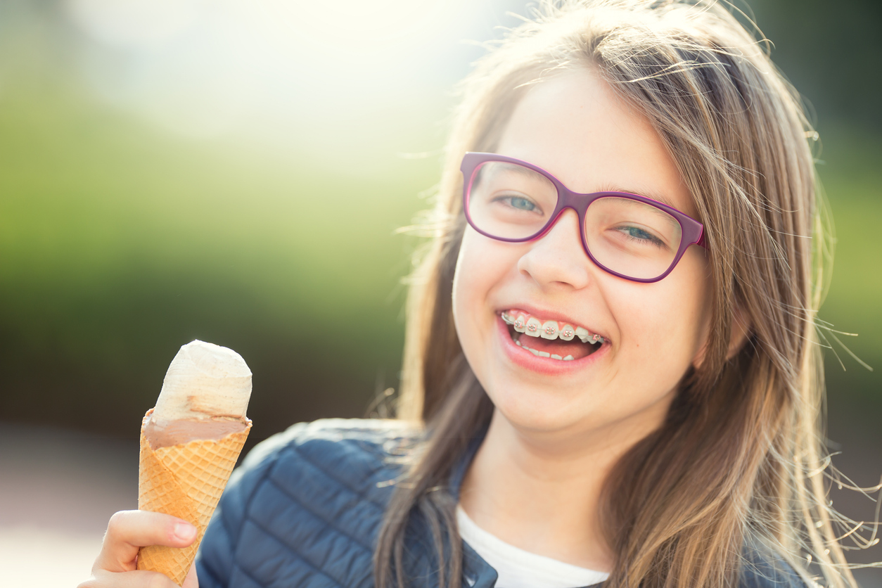 A young girl wearing purple glasses and braces on her teeth, smiling while holding a cone of ice cream or frozen yogurt. 