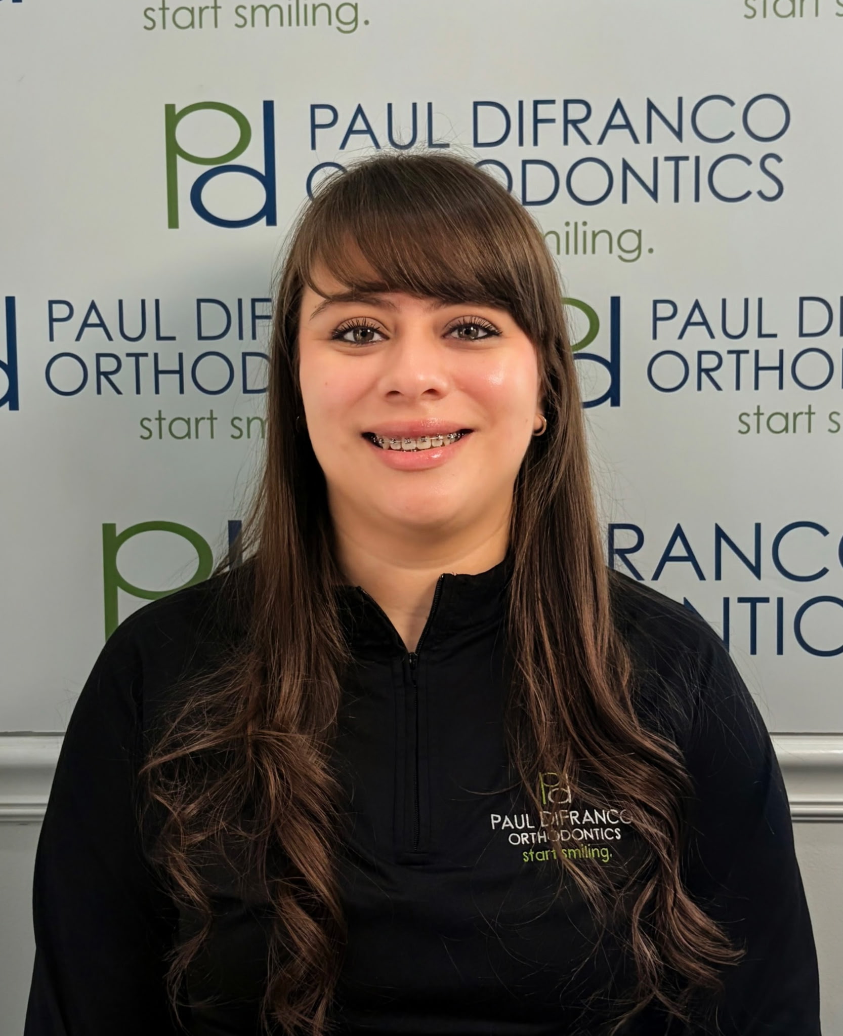 Melissa Martinez, Orthodontic Assistant at Paul DiFranco Orthodontics, wearing a black polo with the logo, smiling in front of a branded backdrop, showcasing her dedication to patient care in Palos Hills, IL.