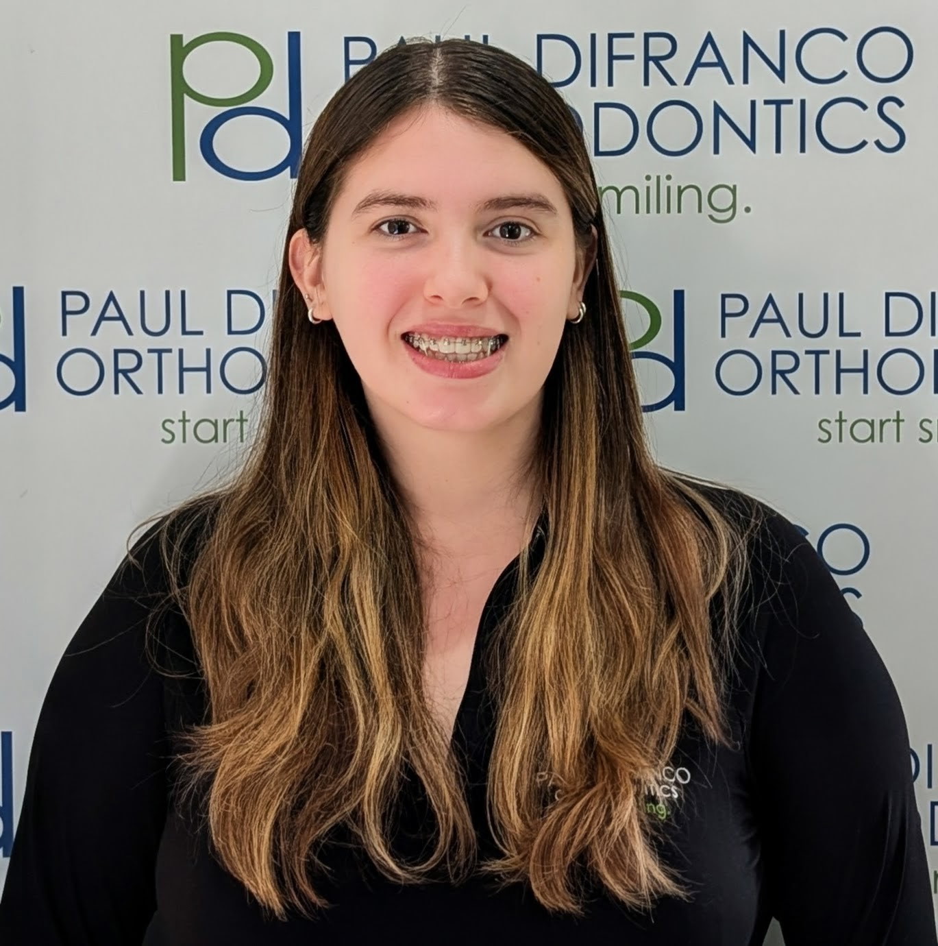 Smiling young woman with braces, wearing a black shirt with the Paul DiFranco Orthodontics logo, against a backdrop featuring the practice name and slogan, representing personalized orthodontic care in Palos Hills, IL.